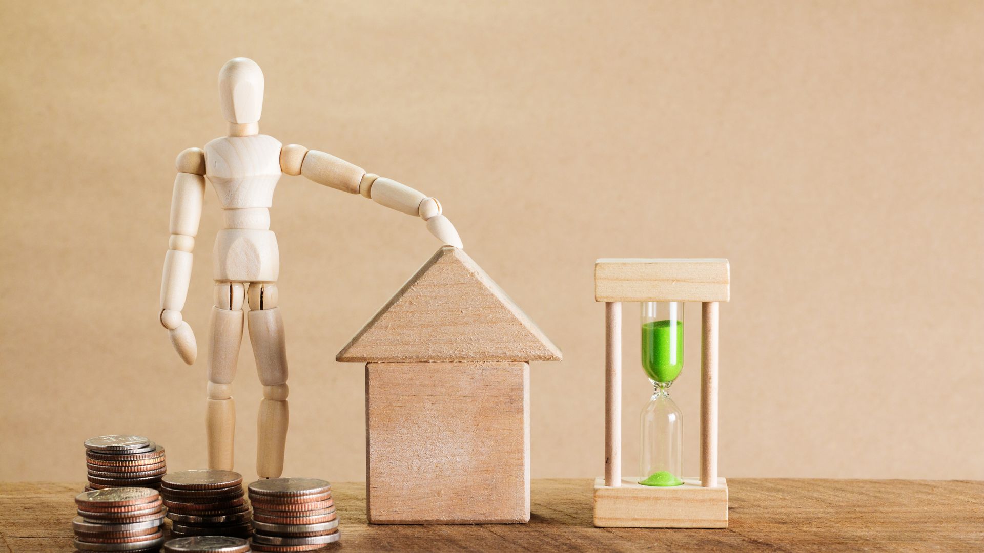 Wooden figure resting a hand on a toy house beside coins and a green hourglass