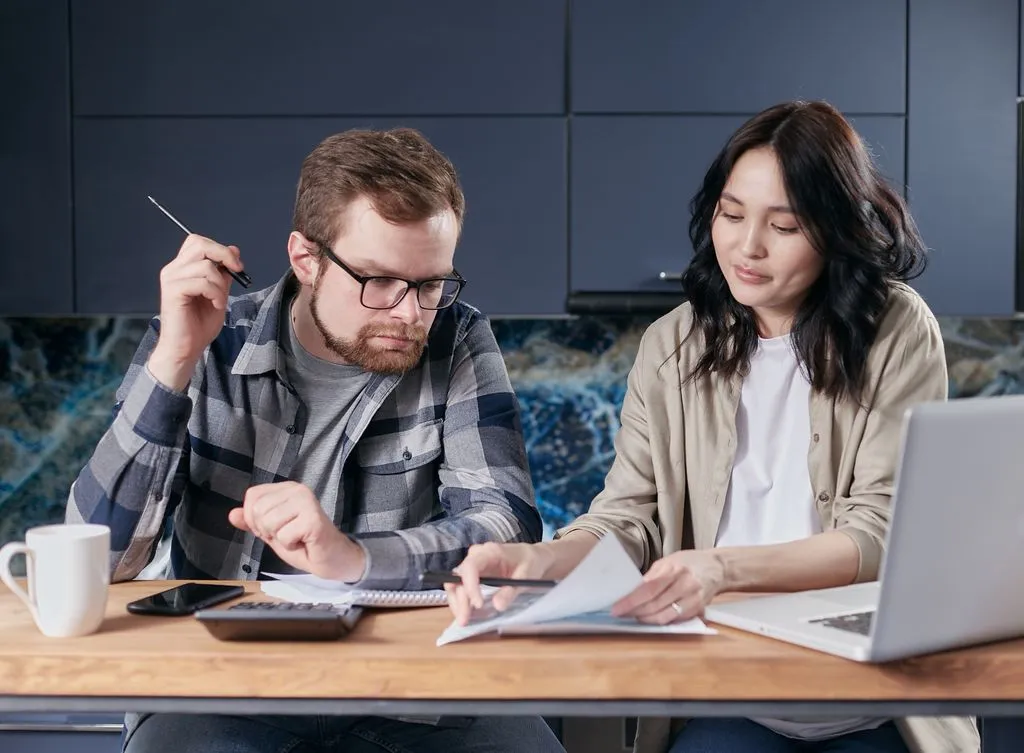 Comparing deals side by side Couple at a kitchen table reviewing documents with a calculator, laptop and mug