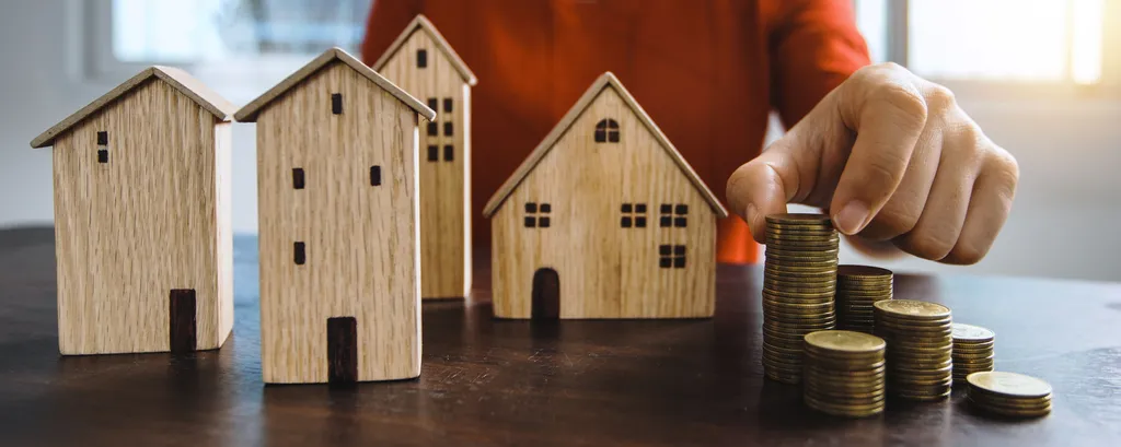 Dłoń układa monety obok drewnianych modeli domów